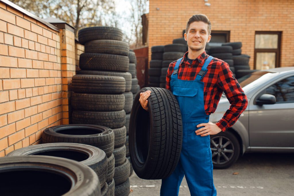 A tire service worker stands holding a tire, with a stack of various used tires in the background.