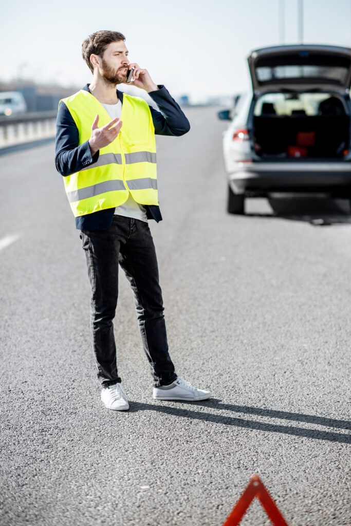 Man calls for roadside assistance after a tire blowout, standing by his car on the highway.