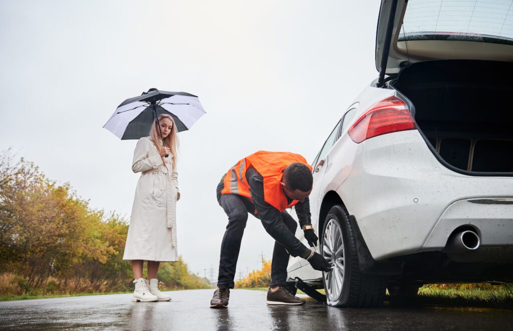 Professional flat tire repair service rescuing a woman stranded with a damaged tire on a rainy day.